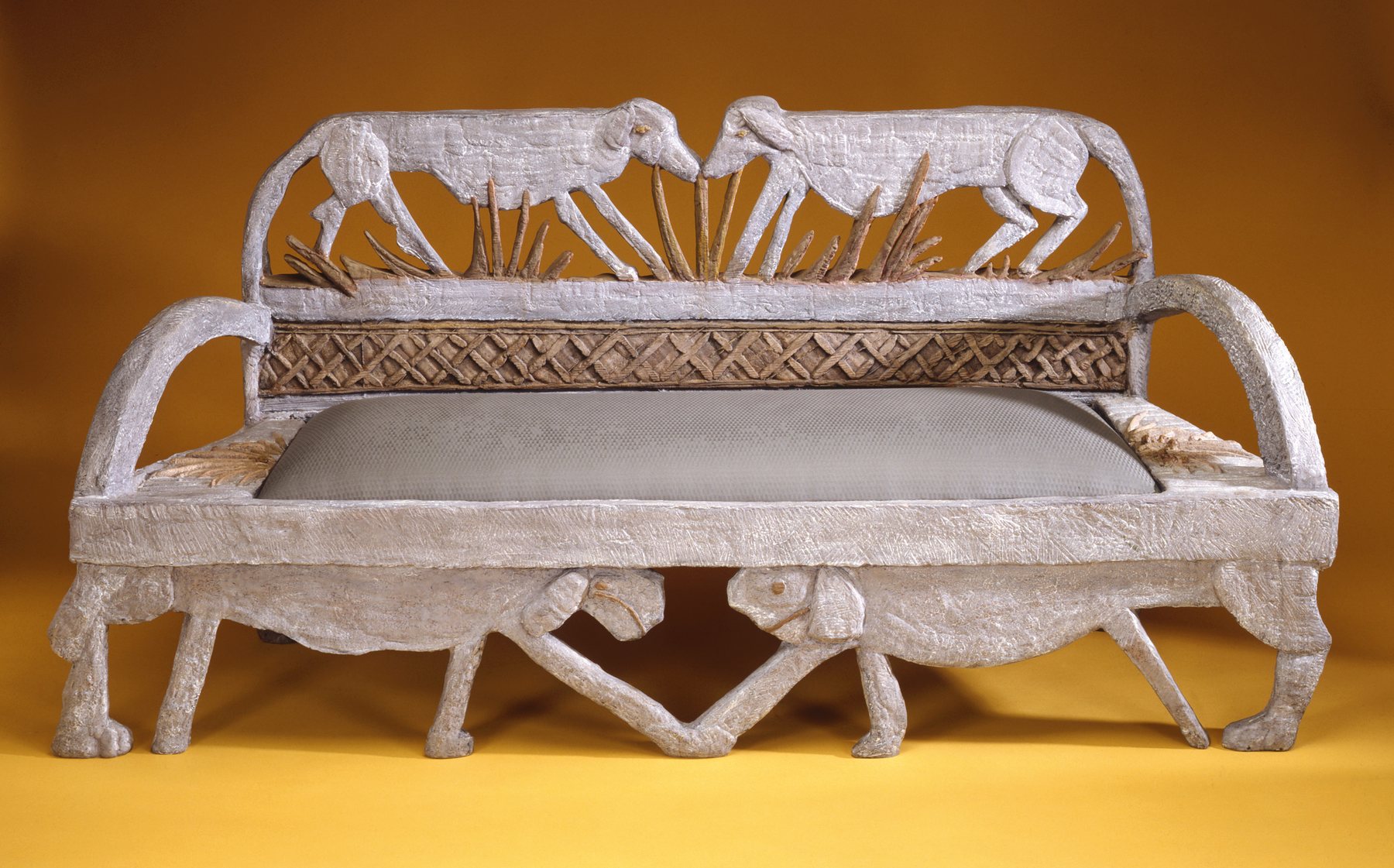 White sculpted bench with an openwork back of elephants, against a yellow background