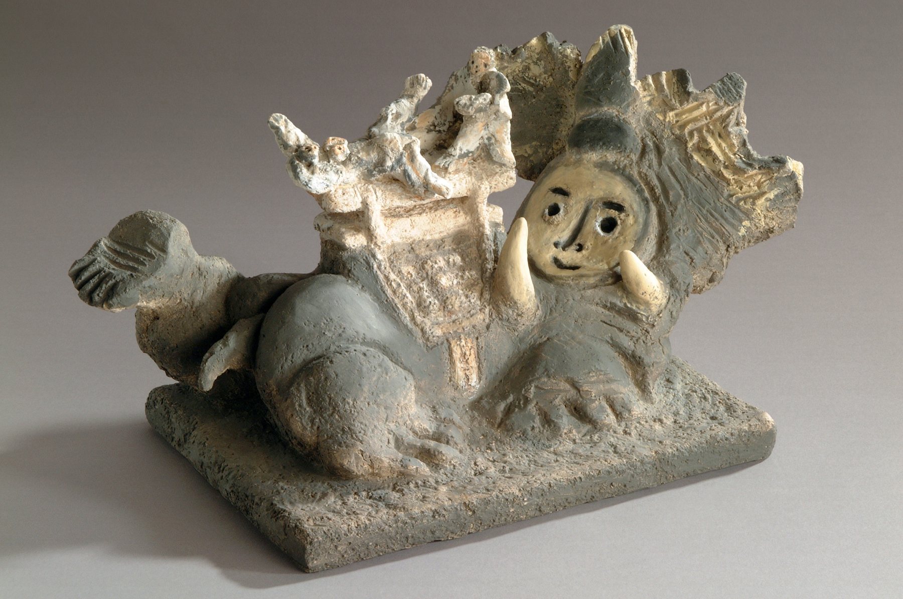 Figura tendida que se sujeta el rostro bajo un elefante dorado, en terracota